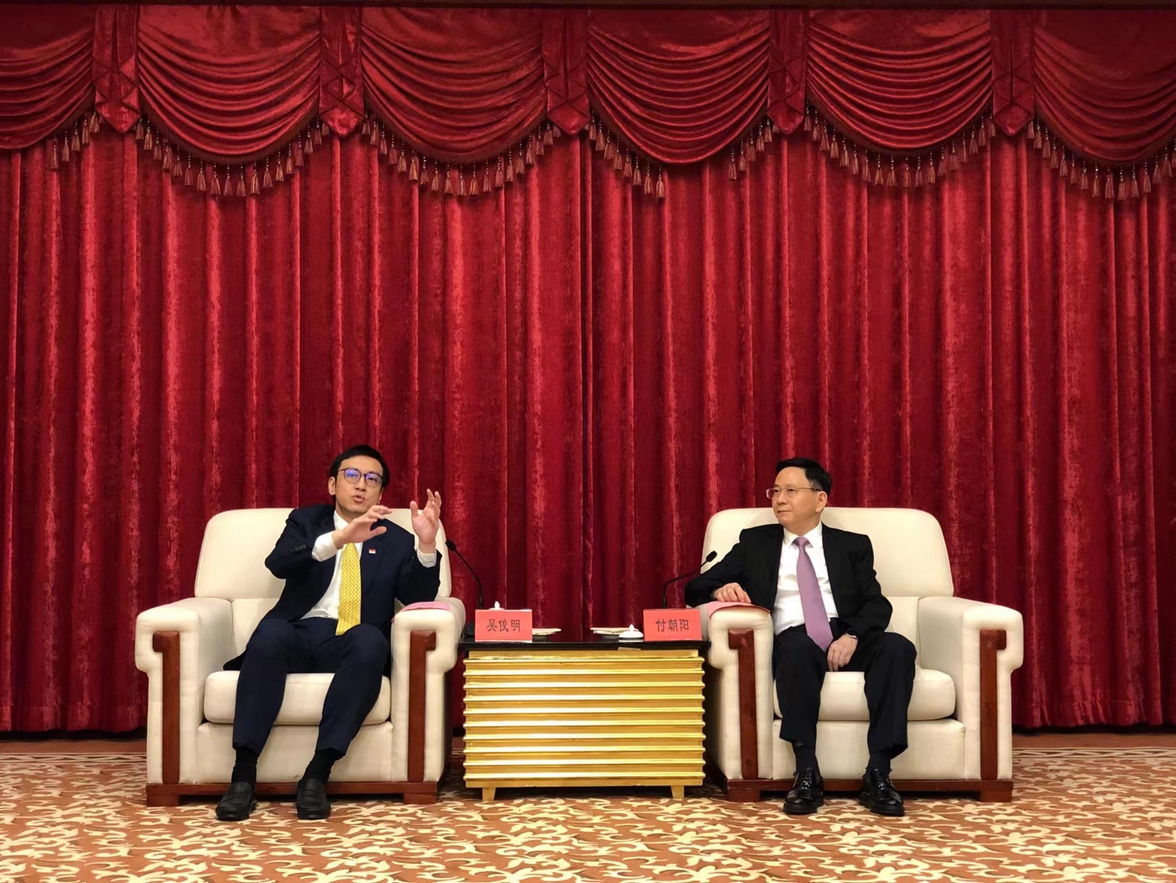 Two men in suits sit in armchairs before red drapes. A table separates them.